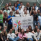 Participants at the General Assembly Prague pose together on a staircase, holding a large event banner inside a modern conference venue.