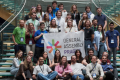 Participants at the General Assembly Prague pose together on a staircase, holding a large event banner inside a modern conference venue.